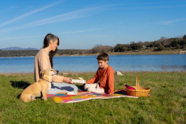 Anne, köpeğiyle göl kenarında piknik yaparken oğluna sandviç veriyor.