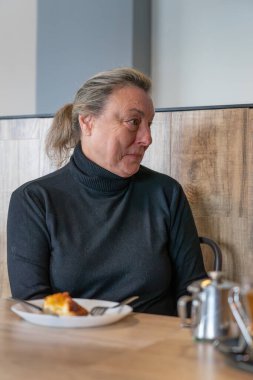 Son sınıf öğrencisi bir kadın, bir kafede sessiz bir kahvaltı yapıyor, düşünceli bir ifadeyle yüzünü çeviriyor.