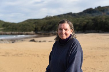 Mutlu, emekli bir kadın güzel bir sahilde duruyor. Arkasında okyanus ve orman var.