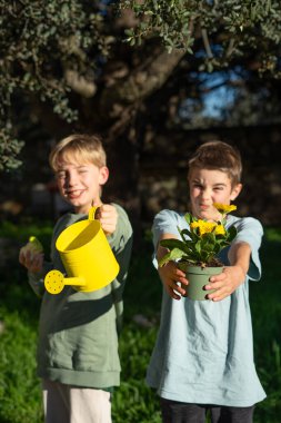 İki kardeş bir çiçek ve bir su bidonu tutuyor güneşli bir günde bir ağacın altında bahçıvanlık yapıyorlar.