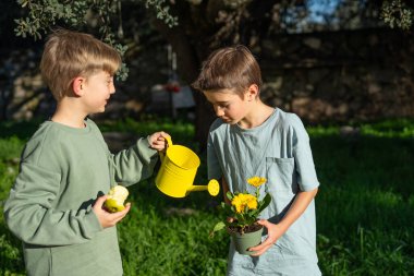 İki genç kardeş, sarı bir su bidonuyla bir saksıda çiçekleri suluyor. Doğa ve bahçe işlerini öğreniyorlar.