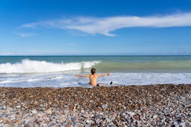 Çocuk okyanus dalgalarına bakan bir çakıl taşı sahilinde oturuyor.