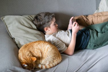 Çocuk kanepede uzanmış, uyuyan bir kedinin yanında telefonla konuşuyor.