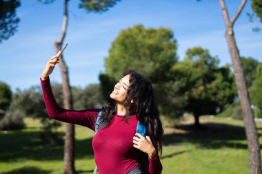 Genç Latin bir kadın güneşli bir günde parkta akıllı telefonuyla selfie çekiyor.