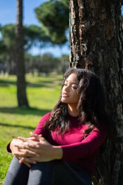 Huzurlu genç Latin kadın güneşli bir parkta ağaçta otururken huzur ve sükunet buluyor.