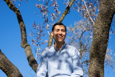 Neşeli genç latin adam Badem ağacı dalları altında mavi gökyüzüne karşı bahar mevsiminin tadını çıkarıyor.