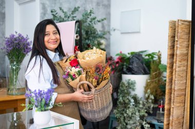 Çiçekçi dükkanında çiçek buketleri ve kurumuş çiçeklerle dolu bir sepet taşıyor.