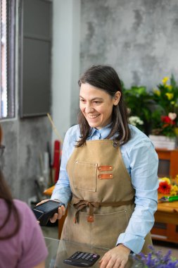 Çiçekçide NFC teknolojisi kullanarak müşteriler akıllı telefonla ödeme yaparken gülen çiçekçi duruş terminalini tutuyor.