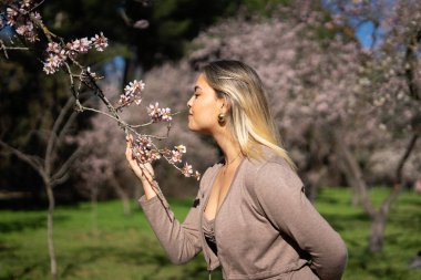 Bahar zamanı güneşli bir bahçede badem çiçeklerinin kokusundan hoşlanan genç bir kadın.