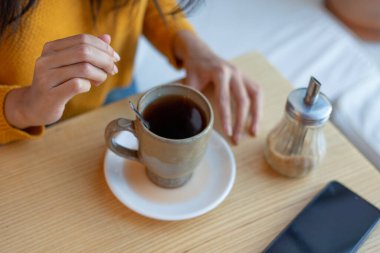 Latin bir kadın kafeterya masasında bir fincan kahveye şeker ekliyor. Yanında da akıllı telefon ve şeker makinesi var.