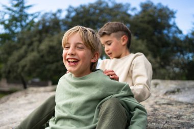 İki genç çocuk bir kayanın üzerinde oturmuş, birlikte gülüyor ve açık havada oynuyorlar.