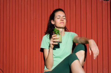 Sporcu kadın gözleri kapalı rahatlıyor ve kırmızı arka planda sağlıklı yeşil meyve suyu tutuyor.