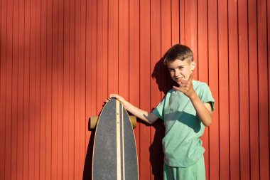 Güneşli bir günde kırmızı ahşap duvarın önünde duran çocuk, uzun tahta bir levha tutuyor ve el işareti yapıyor.