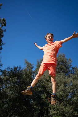 Genç çocuk kolları açık atlarken ve ağaçlarla ve mavi gökyüzüyle parkta bağırırken eğleniyor.