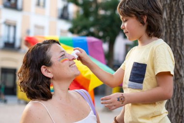 Bir gurur etkinliği sırasında annesinin yüzüne gökkuşağı resmi çizen genç bir çocuk lgbtq + haklarını ve aile sevgisini kutluyor.