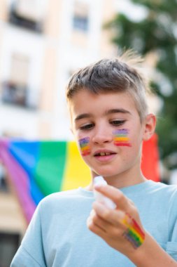 Yüzüne beyaz bir boya çubuğuyla gökkuşağı bayrağı çizen çocuk, çeşitliliği ve kapsamı kutluyor.