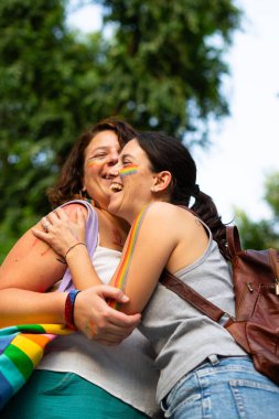 İki kadın kucaklaşıyor ve gülüyor, yüzlerinde gökkuşağı bayraklarıyla bir gurur etkinliğinde aşkı ve eşitliği kutluyorlar.