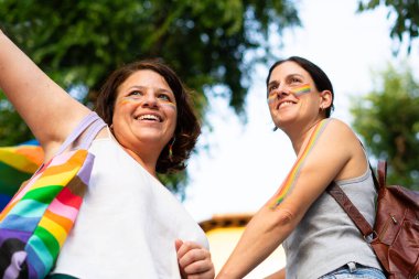 Gökkuşağı makyajlı iki kadın el ele tutuşup eşcinsel onur yürüyüşünü kutluyorlar.