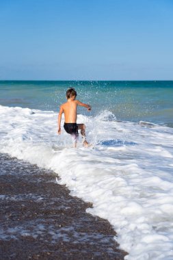 Çocuk sahilde gelen dalgaları tekmeliyor yaz tatilinde eğleniyor.