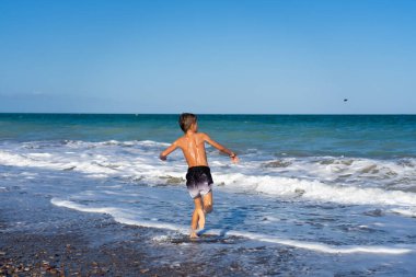 Güneşli bir yaz gününde okyanusa koşan çocuk dalgaların ve kumsalın tadını çıkarıyor.