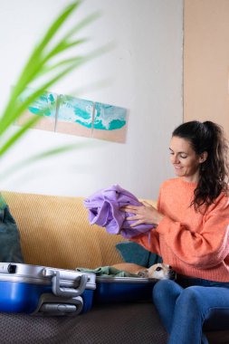 Gülümseyen kadın bavulunu topluyor kanepeye oturuyor ve kıyafetlerini yolculuk için hazırlıyor, köpeği de onu izliyor.