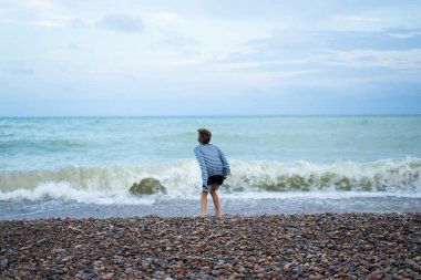 Özgürlüğün tadını çıkaran ve çakıl taşı plajında doğayı keşfeden çocuk, bulutlu bir günde okyanus dalgalarının gücünü gözlemliyor.