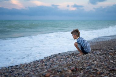 Düşünceli çocuk çakıl taşı plajında çömelmiş, alacakaranlıkta okyanus dalgalarını izliyor, denizin enginliğini düşünüyor.