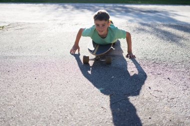 Gülümseyen çocuk uzun bir tahtanın üzerinde uzanıyor ve kendini asfalt yolda parlak güneş ışığında itiyor.
