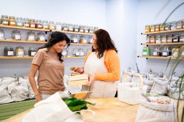 Sıfır atık mağazasında yeniden kullanılabilir poşetler ve cam kavanozlarla zencefil kökü alan bir müşteriye hizmet eden dükkan sahibi.