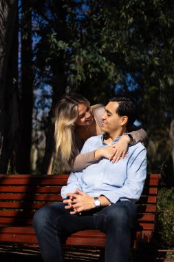 Genç Latin çift parkta güneşli bir günün tadını çıkarıyor, bankta oturuyor ve sarılıyorlar.