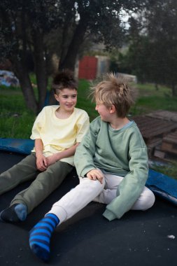 İki genç çocuk trambolinde oturup sohbet ederek eğleniyorlar.