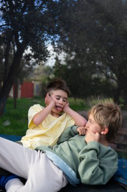 İki kardeş trambolinde uzanıyorlar, aptal suratlar yapıyorlar ve dillerini birbirlerine uzatıyorlar.