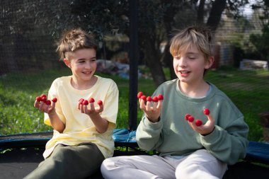 Parmaklarında ahududu gösteren ve bahçede trambolinde otururken gülümseyen iki kardeş.