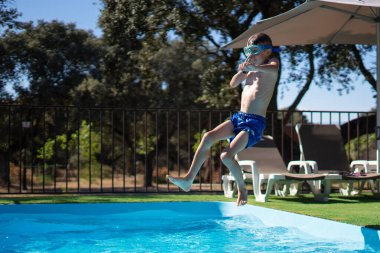 Güneşli bir yaz gününde yüzme gözlüğü takmış bir çocuk yüzme havuzuna atlıyor.