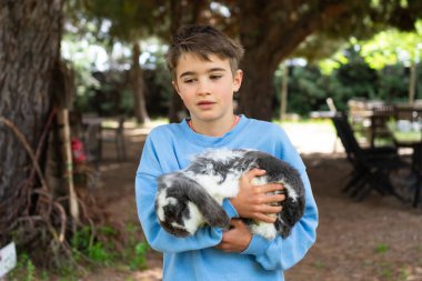 Beyaz çocuk bir çiftlikte tavşanı nazikçe kucaklıyor. Evcil hayvanlara karşı sevgi ve sorumluluk gösteriyor.