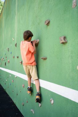 Yapay tırmanma duvarına tırmanma pratiği yapan genç bir çocuk, bir sonraki hamlesine odaklanıyor, gücünü, koordinasyonunu ve problem çözme becerilerini geliştiriyor.