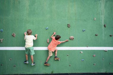 İki genç çocuk yeşil bir tırmanma duvarına tırmanıyor, oyun sahasında eğlenceli ve aktif bir günün tadını çıkarıyorlar.