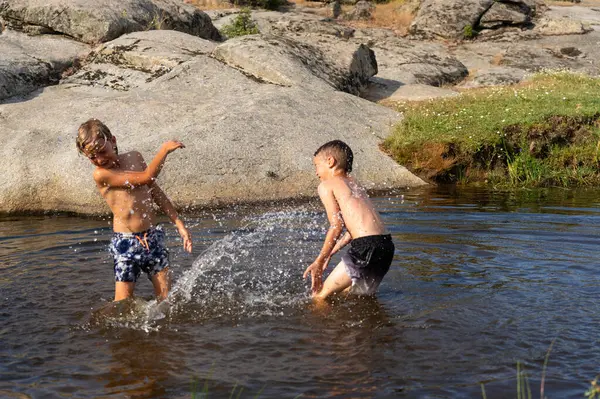 Güneşli bir yaz gününde iki üstsüz çocuk bir nehirde oynuyor ve su sıçratıyor.