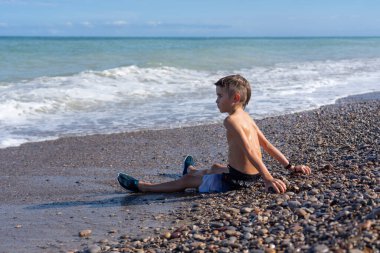 Genç çocuk çakıl taşı plajında oturur, güneşin tadını çıkarır ve dalgaların sahile vuruşunu seyreder.