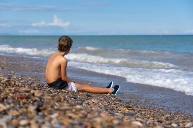 Çakıl taşlı plajda oturan gömleksiz çocuk yaz günü okyanus manzarasının tadını çıkarıyor.