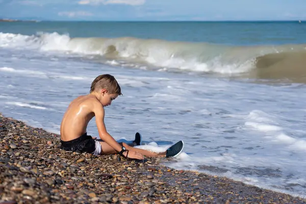 Genç çocuk kumsalda eğleniyor, çakıl taşlarıyla oynuyor güneşli bir günde ayaklarının üzerinde hafif dalgalar süzülürken.