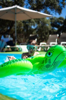 Küçük çocuk yazın tadını çıkarıyor, havuzda şişme kaplumbağayla oynuyor.