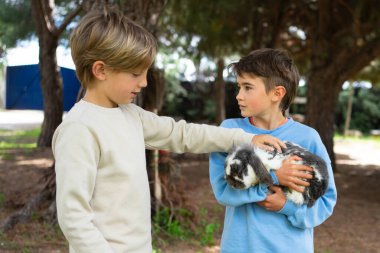 İki çocuk ormanda bir tavşanı okşuyor, hayvanlara ilgi ve şefkat gösteriyorlar.