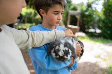 İki çocuk çiftlikte sevimli bir tavşanı okşuyor. Hayvanlara sevgi ve ilgi gösteriyorlar.