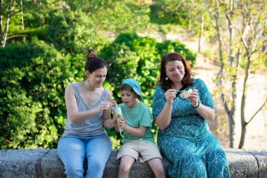 İki kadın ve bir çocuk alçak bir duvarda oturmuş, park ortamında dondurma ve atıştırmalıkların tadını çıkarıyorlar.
