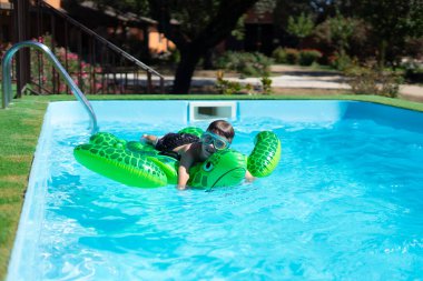 Yüzme gözlüğü takan gülümseyen bir çocuk yaz tatilinde havuzda şişme yeşil bir kaplumbağayla oynuyor.