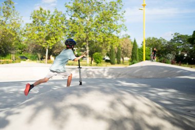 Genç bir çocuk kaykay parkında scooterla geziyor, açık havada egzersiz yapıyor.