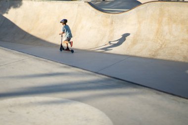 Çocuk beton bir kaykay parkında scooter sürmekten zevk alıyor, güvenlik için kask takıyor.