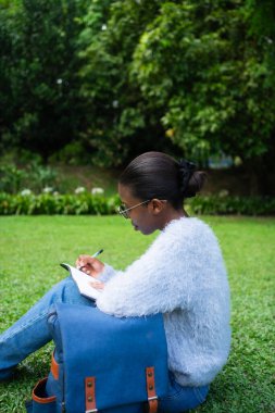 Üniversite öğrencisi dışarıda parkta öğrenirken deftere notlar yazıyor.