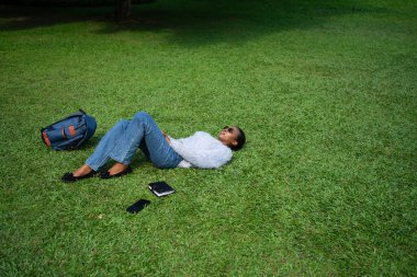 Üniversite öğrencisi bir parkın çimlerinde dinleniyor. Çalışmaya ara veriyor, güneşli günün tadını çıkarıyor.